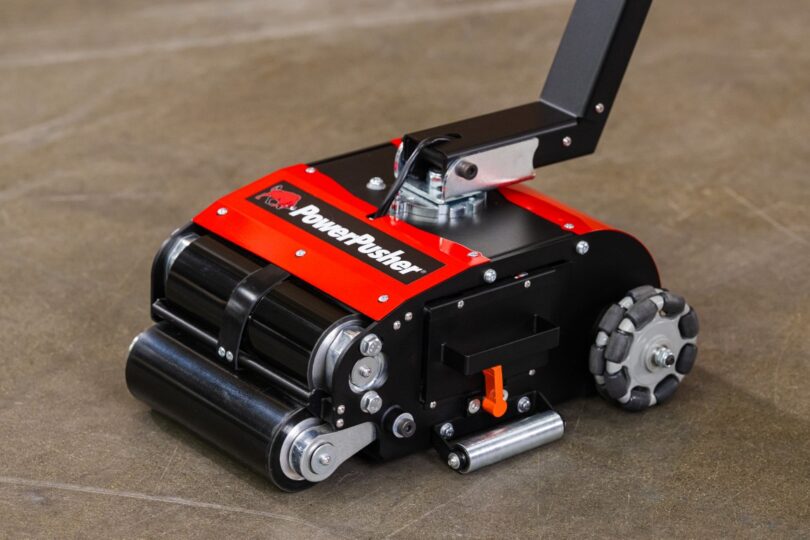PowerPusher electric paper roll mover with red and black housing, drive rollers, and omnidirectional wheel on warehouse floor